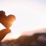 Hand holding a small heart-shaped object against the backdrop of a bright sunset, symbolizing love, gratitude, and reflection.