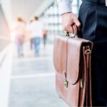Close-up of a man in business attire holding a brown leather briefcase while standing in a bright walkway, with people walking in the background.