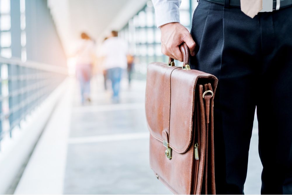 Close-up of a man in business attire holding a brown leather briefcase while standing in a bright walkway, with people walking in the background.