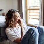 A woman sitting on the floor beside a bed, looking out a window with a thoughtful and anxious expression, symbolizing emotional distress or anxiety.