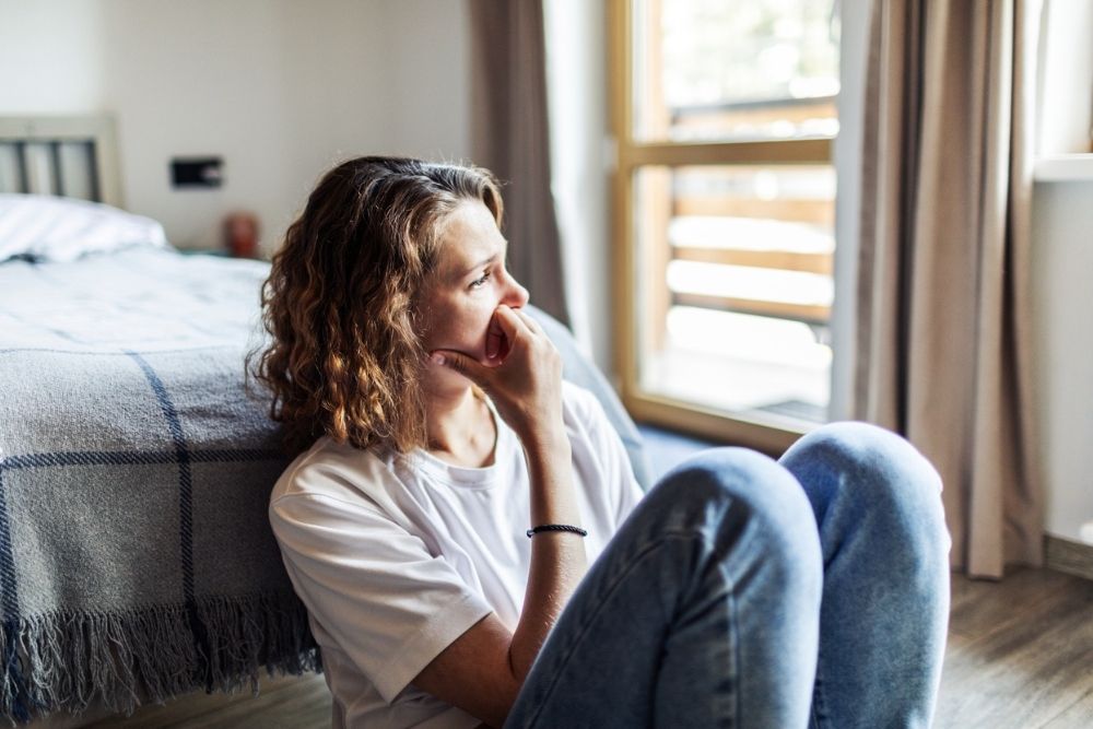 A woman sitting on the floor beside a bed, looking out a window with a thoughtful and anxious expression, symbolizing emotional distress or anxiety.