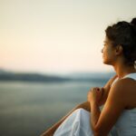 A woman sits outdoors at sunset, gazing thoughtfully over a calm body of water. She rests her arms on her knees and appears peaceful and reflective.