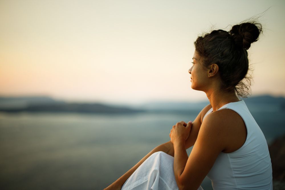 A woman sits outdoors at sunset, gazing thoughtfully over a calm body of water. She rests her arms on her knees and appears peaceful and reflective.