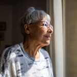 An older woman with short gray hair and glasses gazes thoughtfully out a window, with soft light illuminating her face, suggesting reflection or hope.