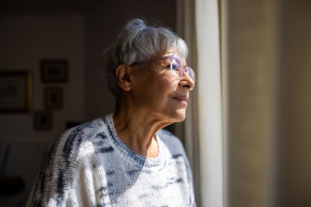 An older woman with short gray hair and glasses gazes thoughtfully out a window, with soft light illuminating her face, suggesting reflection or hope.