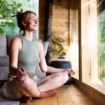 Young woman sitting cross-legged indoors by a large window, smiling peacefully while practicing yoga and meditation.