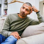 Man sitting on a couch looking worried and deep in thought, suggesting stress or anxiety.
