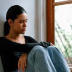 Young woman sitting by a window with a thoughtful, sad expression, hugging her knees as she looks outside.