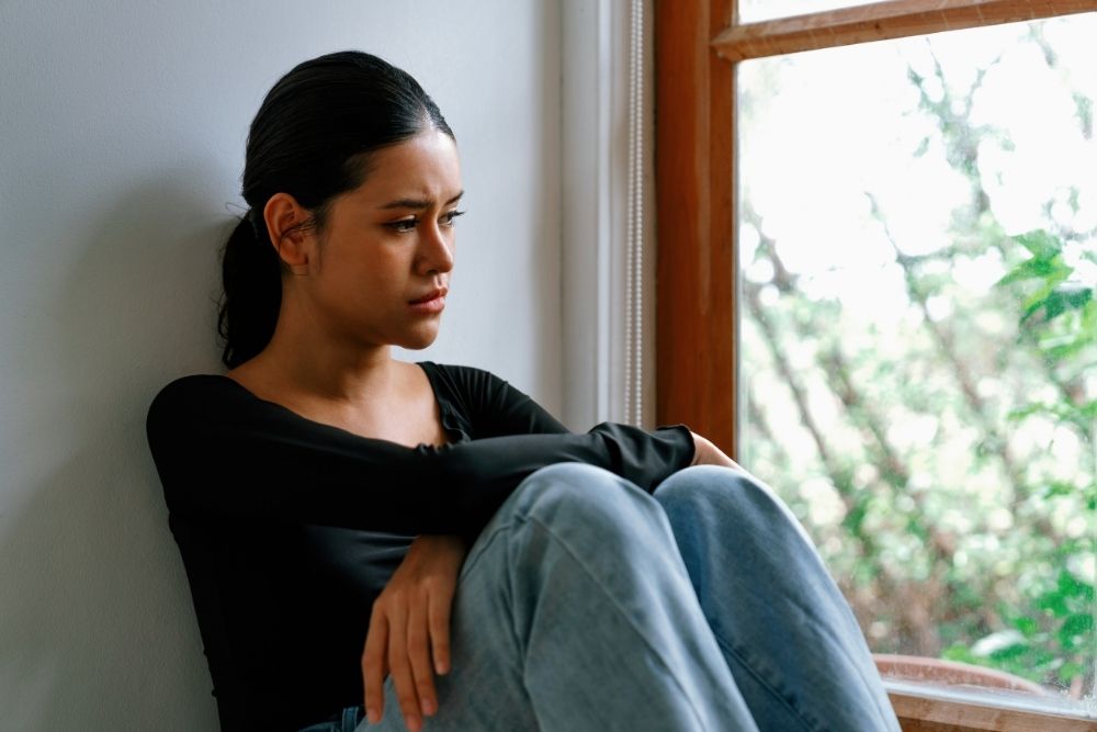 Young woman sitting by a window with a thoughtful, sad expression, hugging her knees as she looks outside.