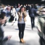 A woman stands in a crowded area covering her face with her hands, while the blurred motion of people around her suggests panic or overwhelm.