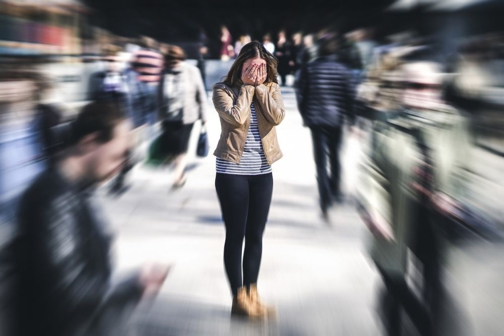 A woman stands in a crowded area covering her face with her hands, while the blurred motion of people around her suggests panic or overwhelm.