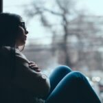 A teenage girl sits by a window with her knees pulled close, looking out pensively, symbolizing sadness, worry, or adolescent depression.