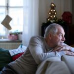 An older man sits on a couch during the holiday season, leaning forward with his arms resting on the back of the sofa as he gazes thoughtfully out a window.