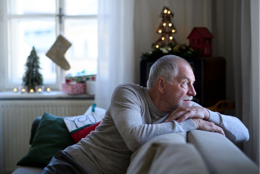 An older man sits on a couch during the holiday season, leaning forward with his arms resting on the back of the sofa as he gazes thoughtfully out a window.
