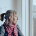 A woman with a ponytail and pink scarf sits indoors by a large window, looking out thoughtfully on a cold, overcast day, conveying a sense of sadness or seasonal depression.