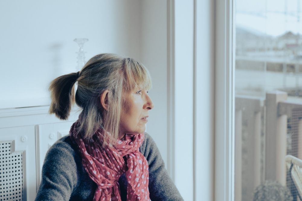 A woman with a ponytail and pink scarf sits indoors by a large window, looking out thoughtfully on a cold, overcast day, conveying a sense of sadness or seasonal depression.