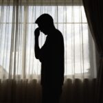 A silhouetted person stands by a window with head bowed and hand to their forehead, expressing emotional exhaustion or distress.