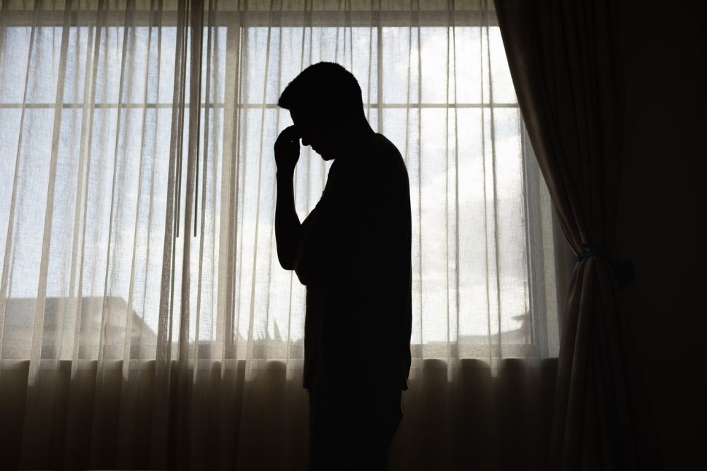 A silhouetted person stands by a window with head bowed and hand to their forehead, expressing emotional exhaustion or distress.