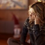 A woman sitting indoors during the holidays, resting her head on her hand and appearing stressed or overwhelmed, with Christmas lights softly glowing behind her.