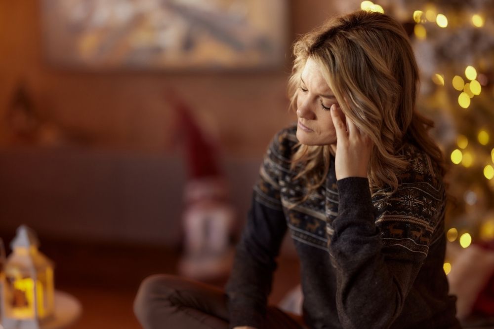 A woman sitting indoors during the holidays, resting her head on her hand and appearing stressed or overwhelmed, with Christmas lights softly glowing behind her.