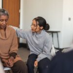 Two women seated in a group setting, one offering comfort and support to the other during a group therapy session.