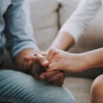 Two people sitting close together, holding hands in a supportive gesture, representing care, connection, and emotional support.