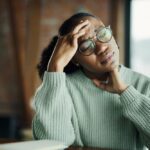 A woman with glasses sits at a table, resting her head in her hands, appearing deep in thought or distress.