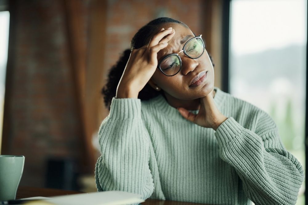 A woman with glasses sits at a table, resting her head in her hands, appearing deep in thought or distress.