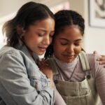 Understanding Borderline Personality Disorder - Two women sit on a couch, embracing each other in support. The setting is a cozy, bright living room. They both appear emotional, conveying comfort and empathy.