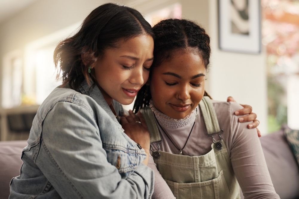 Understanding Borderline Personality Disorder - Two women sit on a couch, embracing each other in support. The setting is a cozy, bright living room. They both appear emotional, conveying comfort and empathy.