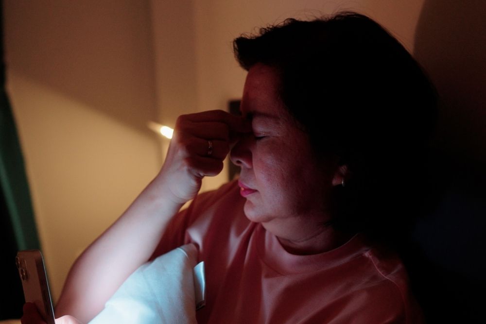 Willow Creek - What Is Prolonged Grief Disorder. A woman in a poorly lit room pinches the crane of her nose. She has her eyes close and looks upset.